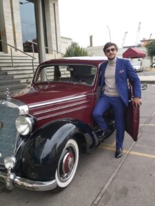 Novio posando junto a auto clásico rojo con traje azul a la medida.