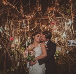 Boda nocturna bajo luces de árbol, novio besando a la novia con traje oscuro.