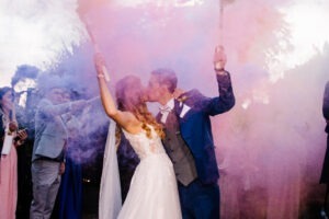 Beso de novios con humo rosa, él lleva un traje azul oscuro.