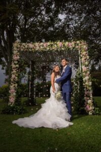 Novios bajo arco floral, él viste un traje azul sartorial perfecto.