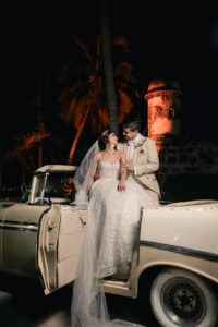 Pareja junto a auto clásico blanco, novio en traje beige a la medida.