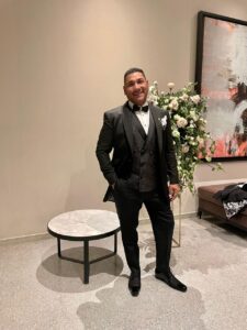 Caballero de pie en interior vistiendo un traje negro clásico impecable.